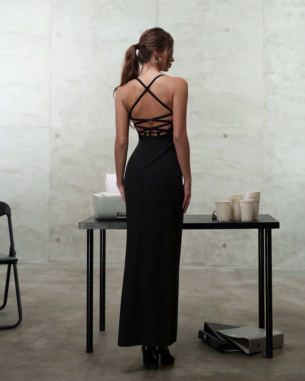 Woman in a black dress with criss-cross straps standing in a minimalistic room.