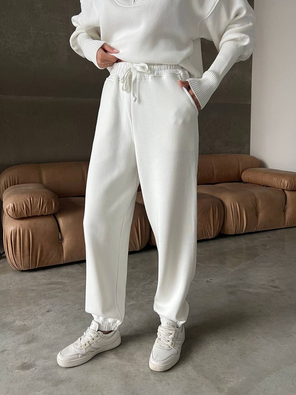 Person wearing a white outfit with jogger pants in a room with brown sofas.