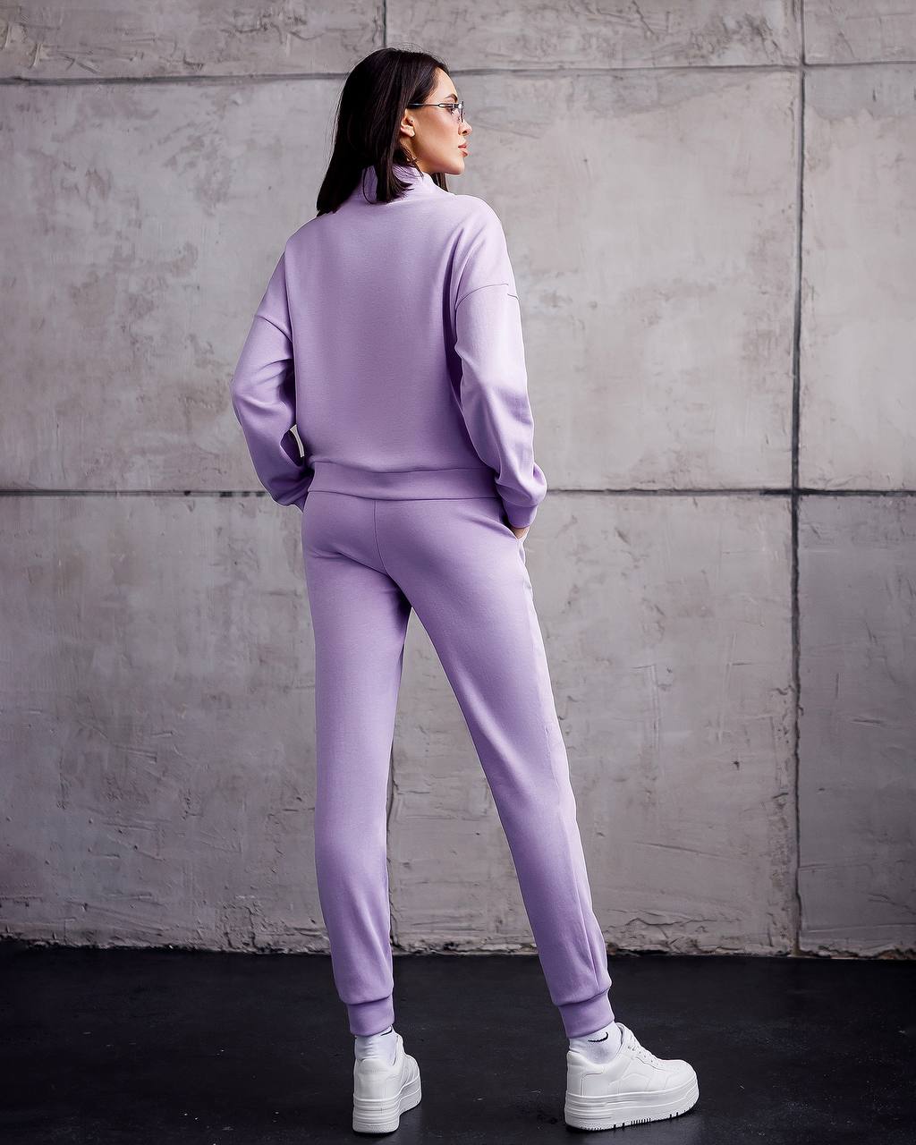 Person wearing a lavender tracksuit standing against a gray concrete wall.