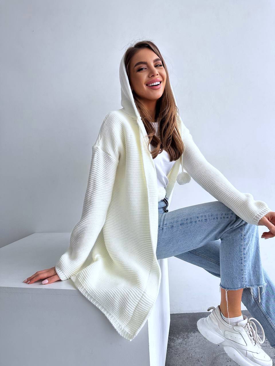 Woman wearing a white hoodie and jeans sitting on a white surface with a light gray background