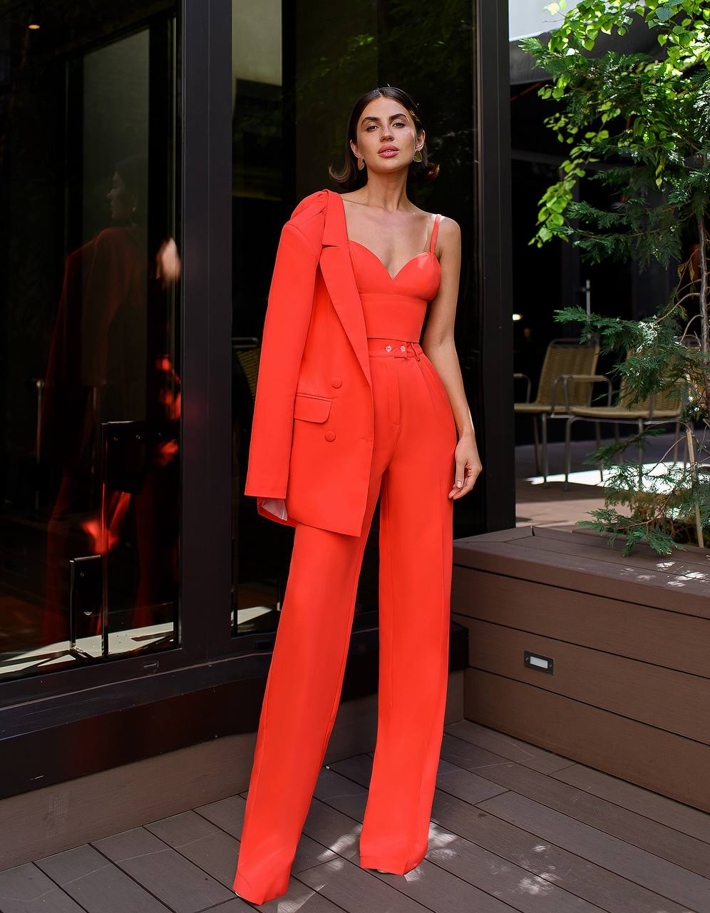Woman in a bright red outfit standing on a wooden deck with a building and plants in the background.