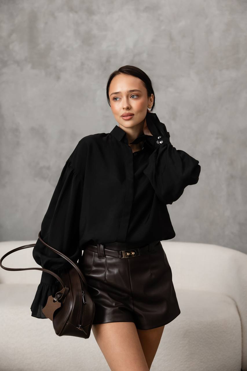 Woman in black outfit with leather shorts holding a brown bag against a gray background