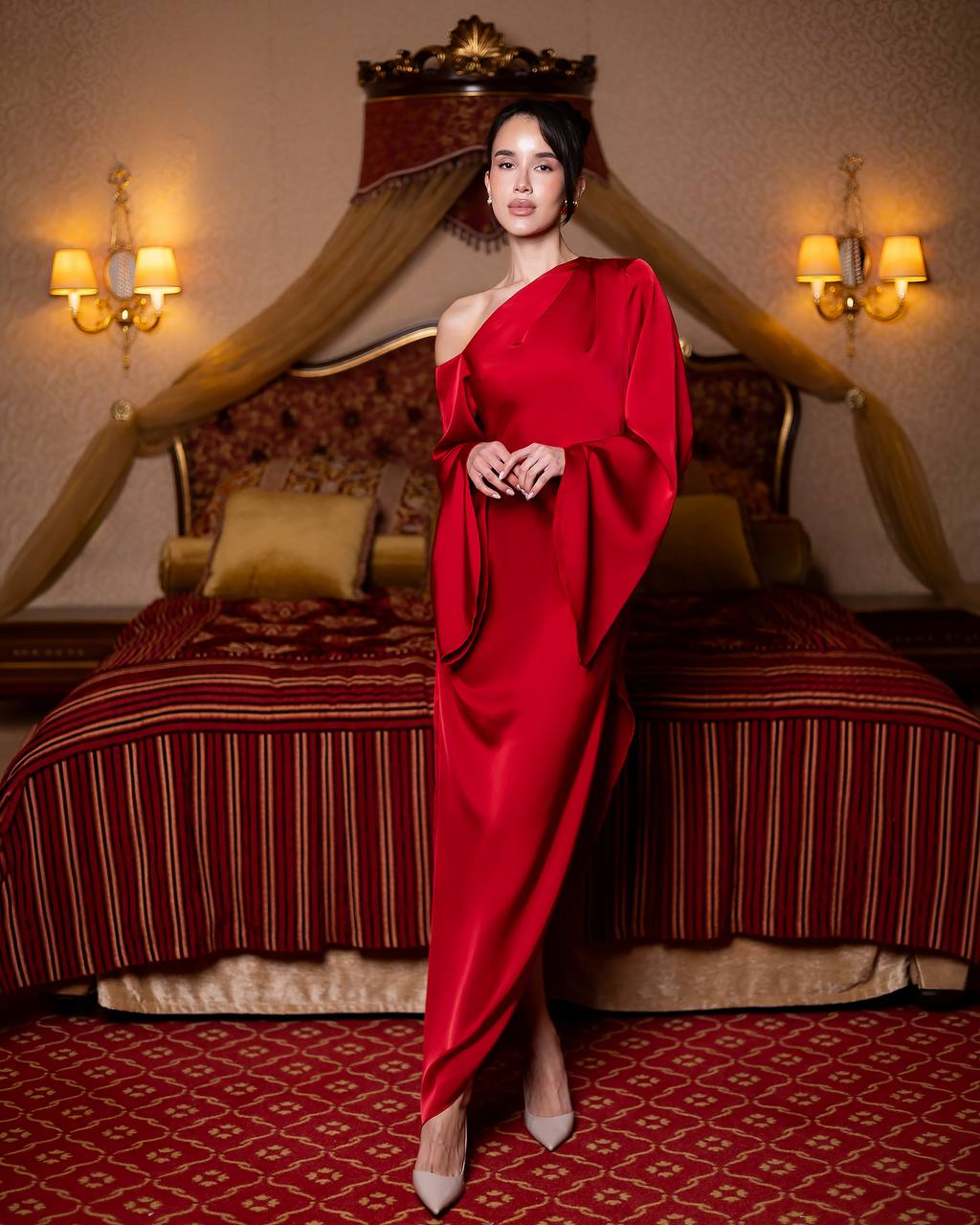A woman stands in a luxurious bedroom wearing a floor-length, one-shoulder red gown with draped sleeves and nude heels. The room features a bed with red and gold striped bedding, a canopy headboard, and warm wall sconces.