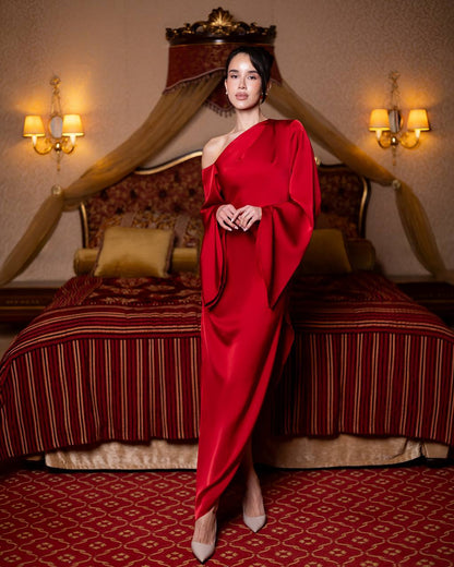 A woman stands in a luxurious bedroom wearing a floor-length, one-shoulder red gown with draped sleeves and nude heels. The room features a bed with red and gold striped bedding, a canopy headboard, and warm wall sconces.