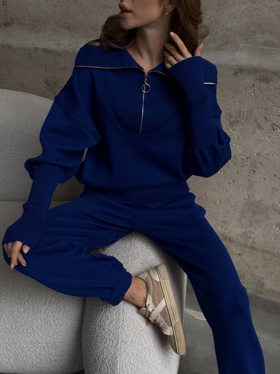 Person wearing a blue tracksuit sitting on a gray chair against a neutral wall.