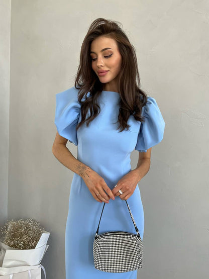 Woman wearing a light blue dress with puffed sleeves, holding a black and white checkered handbag against a plain background.