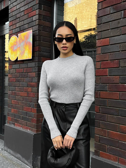 Woman in a gray sweater and black leather skirt standing against a brick wall.