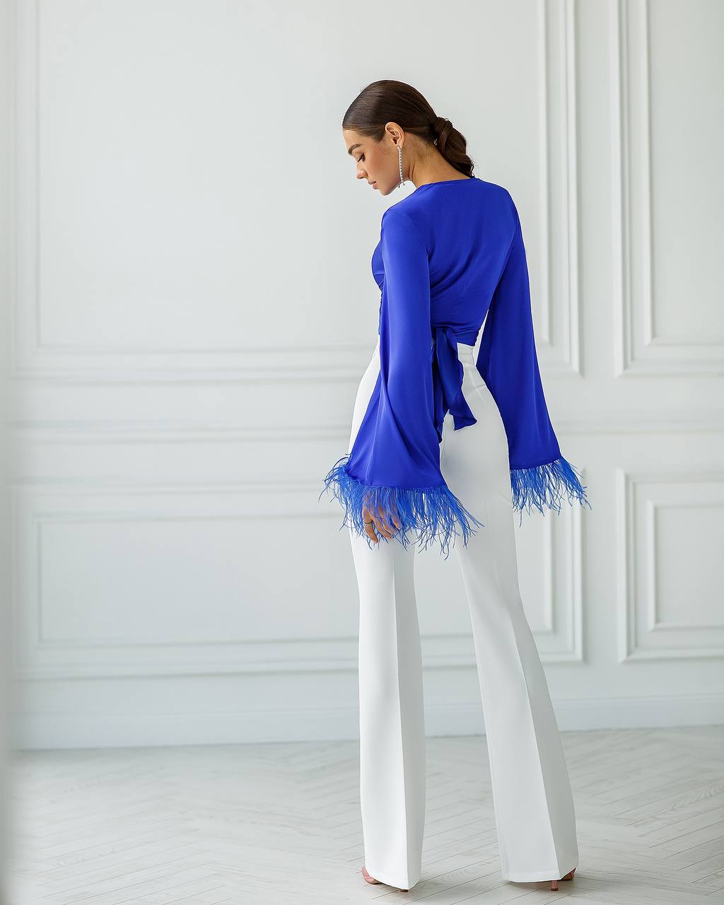 Woman wearing a blue top with feather details and white pants against a white wall.