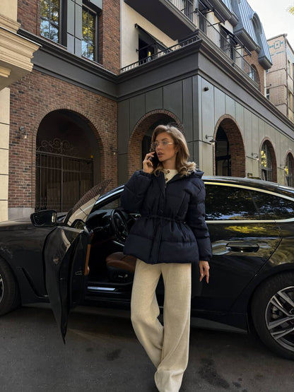 Woman in a black coat and beige pants walking towards a black car on a city street.