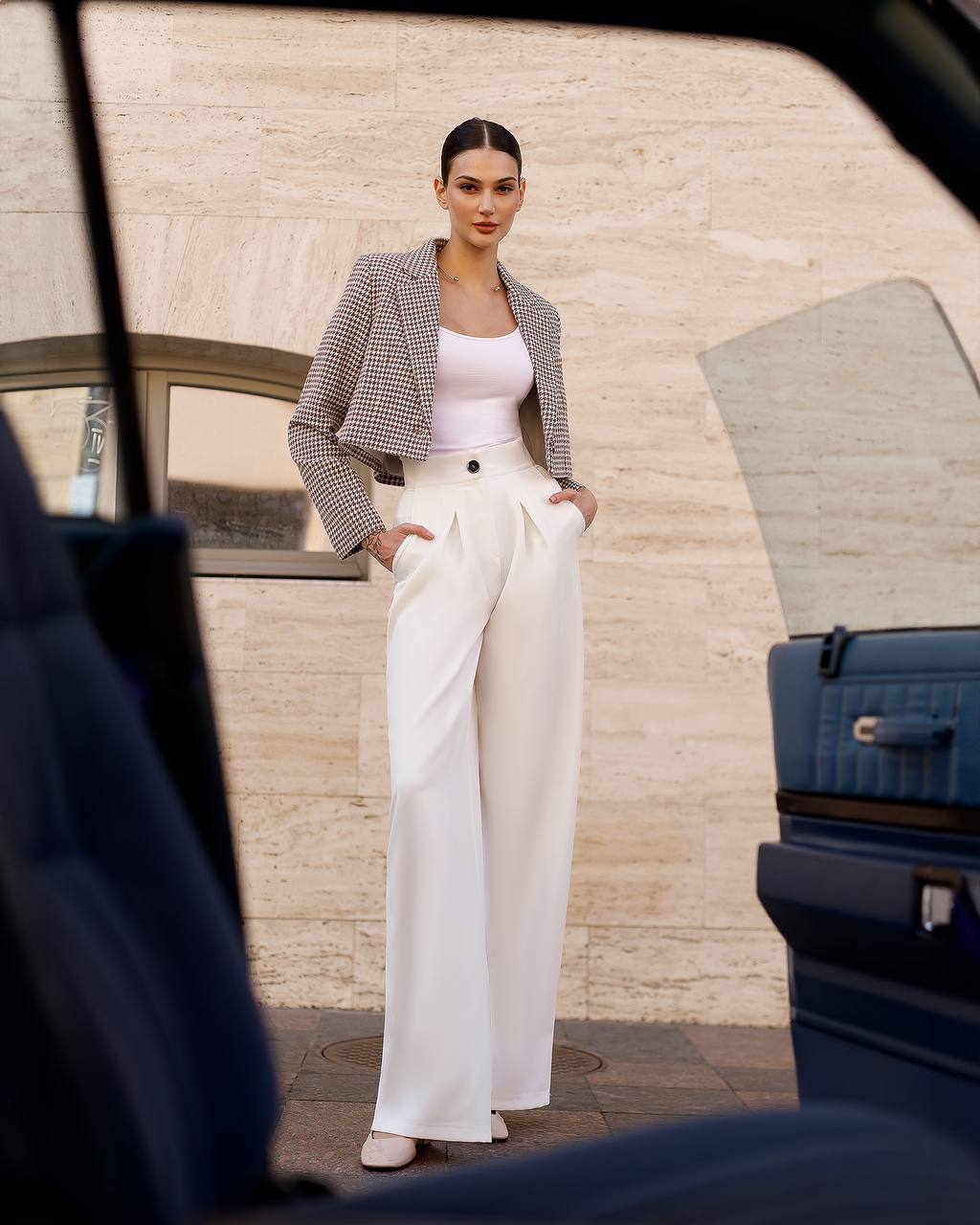 Woman in a stylish outfit standing next to a car with a suitcase.