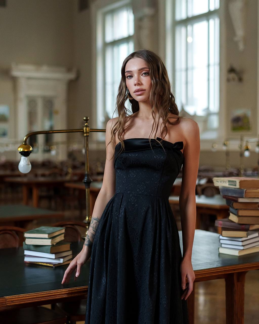 Woman in a black dress standing in a room with books and a lamp.
