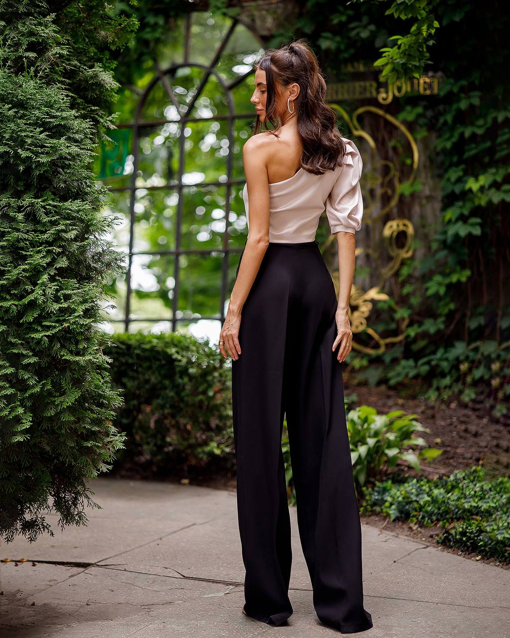 Woman in a white top and black pants standing outdoors with greenery and a decorative archway in the background.