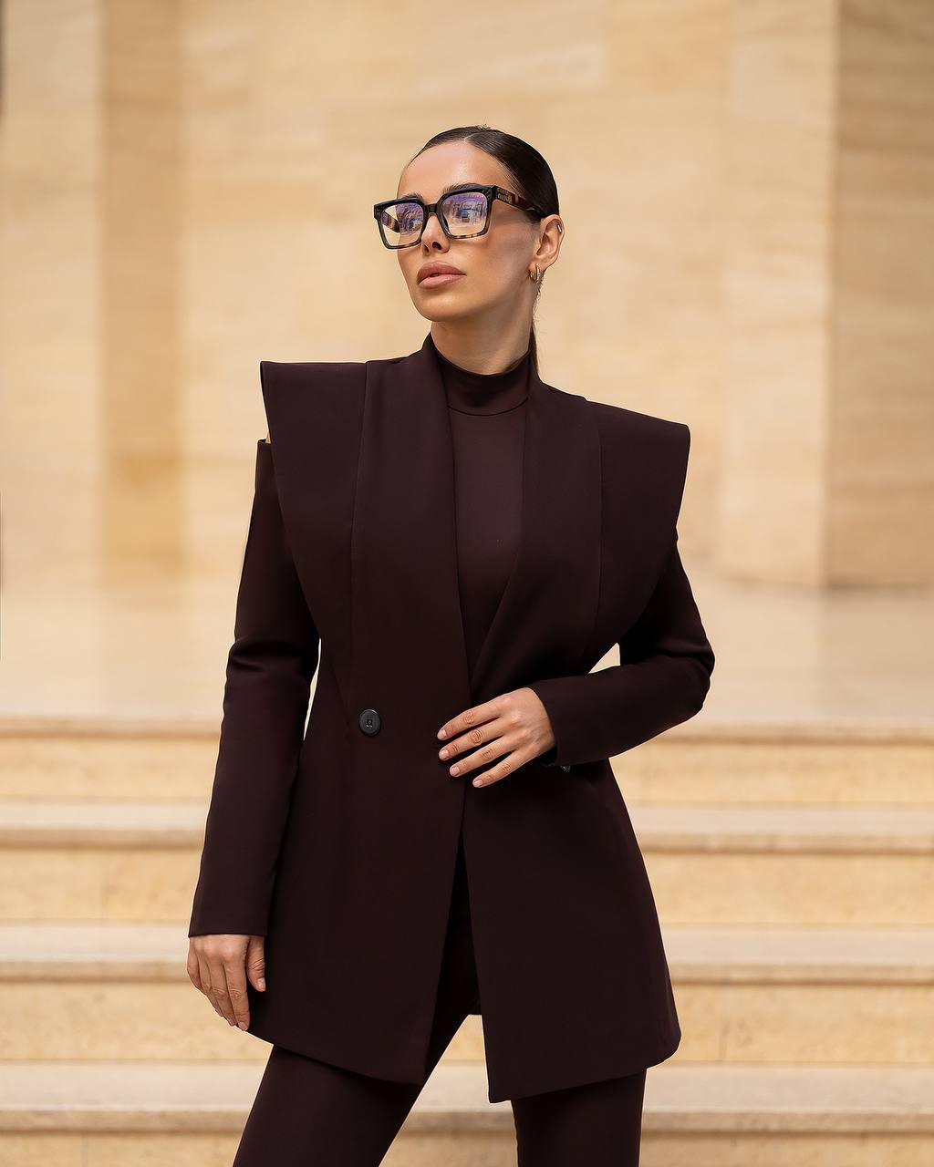 A woman wearing a dark plum-brown structured blazer with dramatic shoulder peaks, a matching mock neck top, and trousers, styled with chunky black glasses, standing on stone steps.