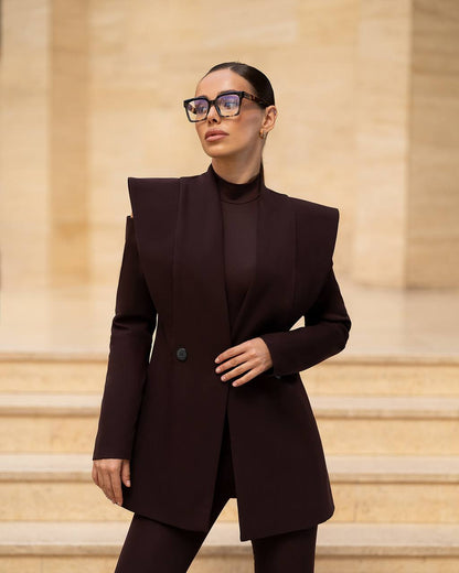A woman wearing a dark plum-brown structured blazer with dramatic shoulder peaks, a matching mock neck top, and trousers, styled with chunky black glasses, standing on stone steps.