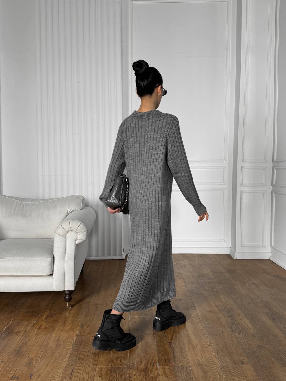 Woman in a gray dress standing in a room with white walls and a gray sofa.