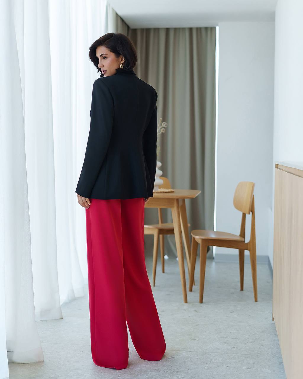 Woman in a black blazer and red pants standing in a modern room with wooden furniture.