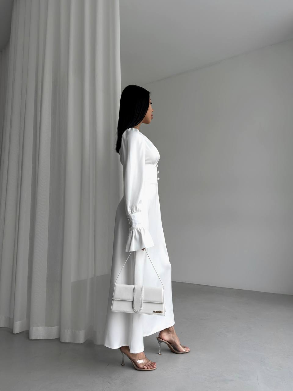 Woman in a white outfit standing in a minimalistic room with gray curtains.