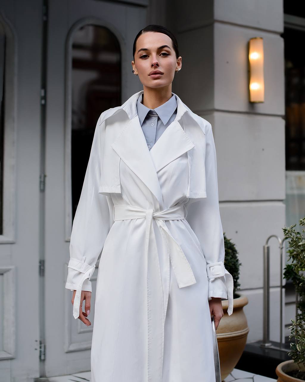 Woman wearing a white trench coat standing outdoors near a building.