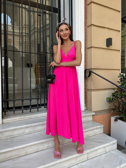 A woman stands on outdoor stone steps wearing a bright hot pink sleeveless maxi dress with a deep V-neckline. She is holding a small black handbag and wearing pink platform sandals, posing in front of a beige building with a black metal gate.