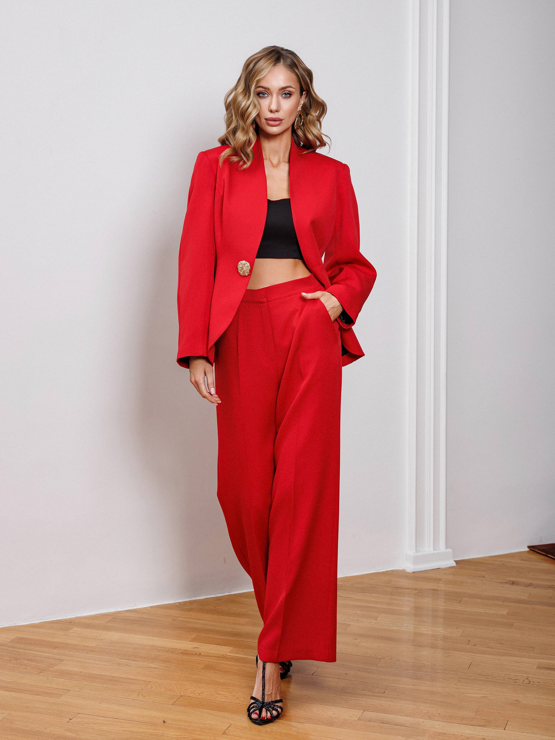 Woman wearing a red suit standing in a room with wooden floor and white walls.