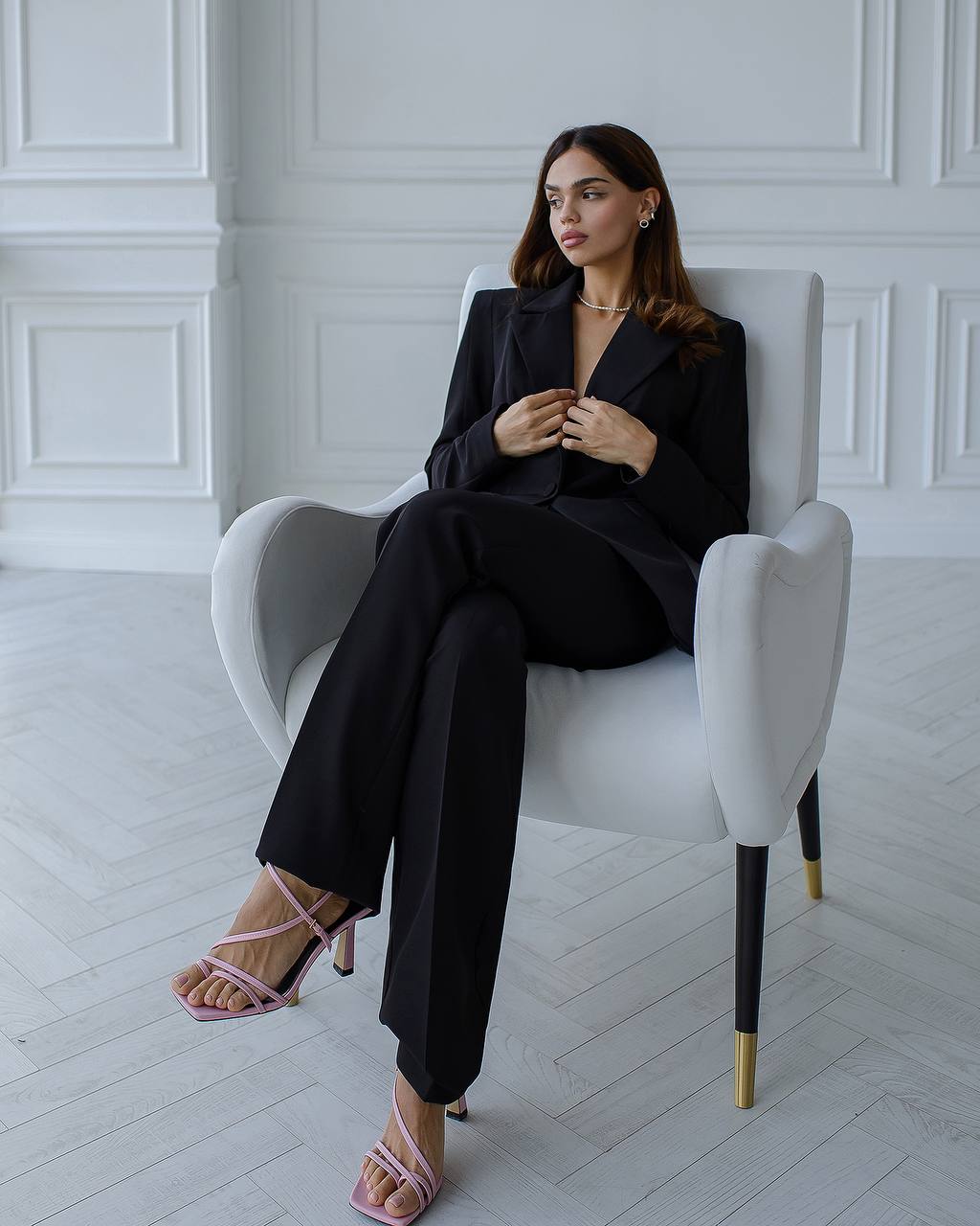 Woman in a black suit sitting on a white chair in a minimalistic room.