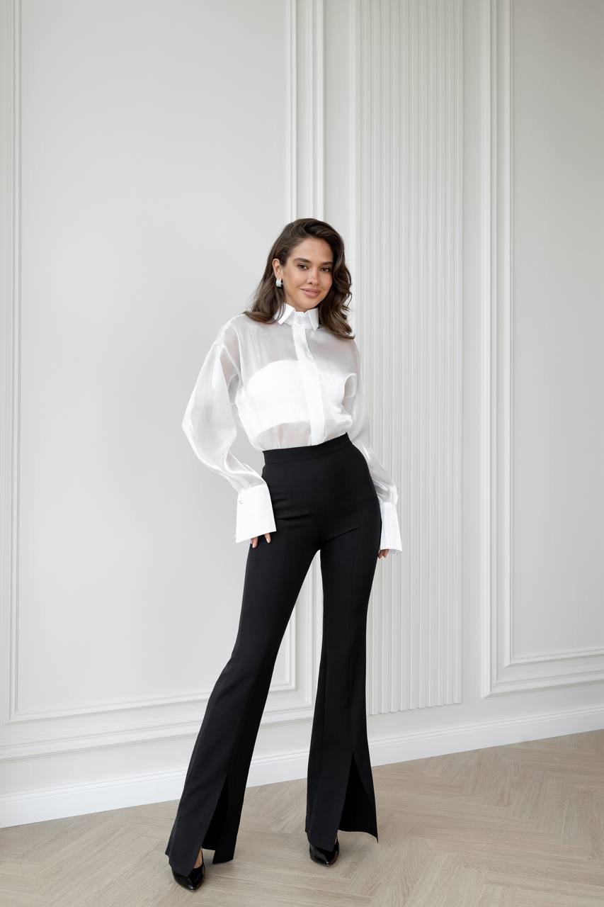 Woman wearing a white blouse and black pants against a white paneled wall.