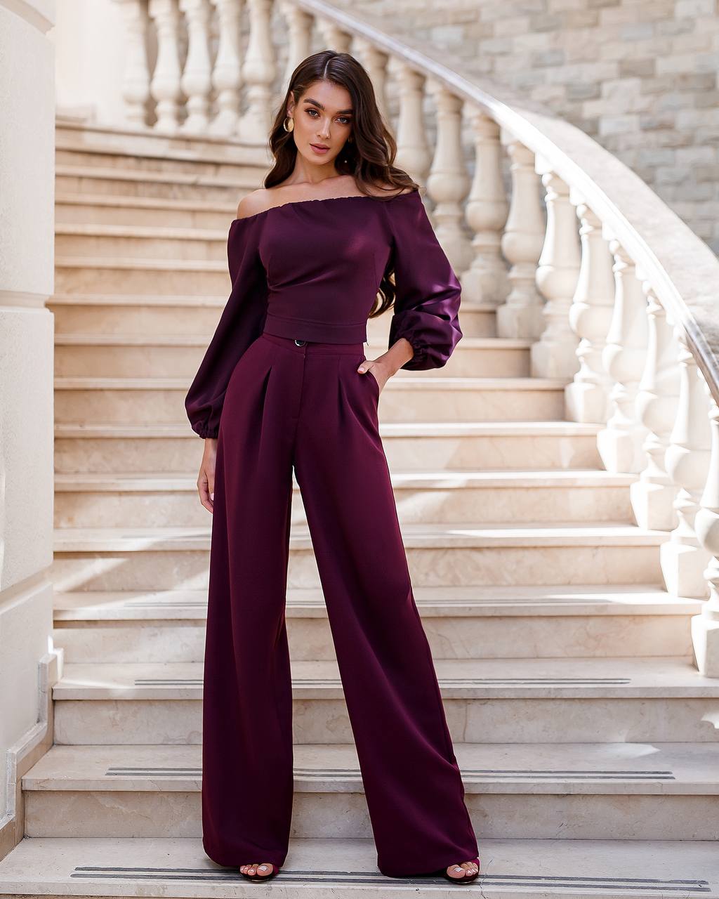 Woman in a burgundy off-shoulder jumpsuit standing on a staircase.