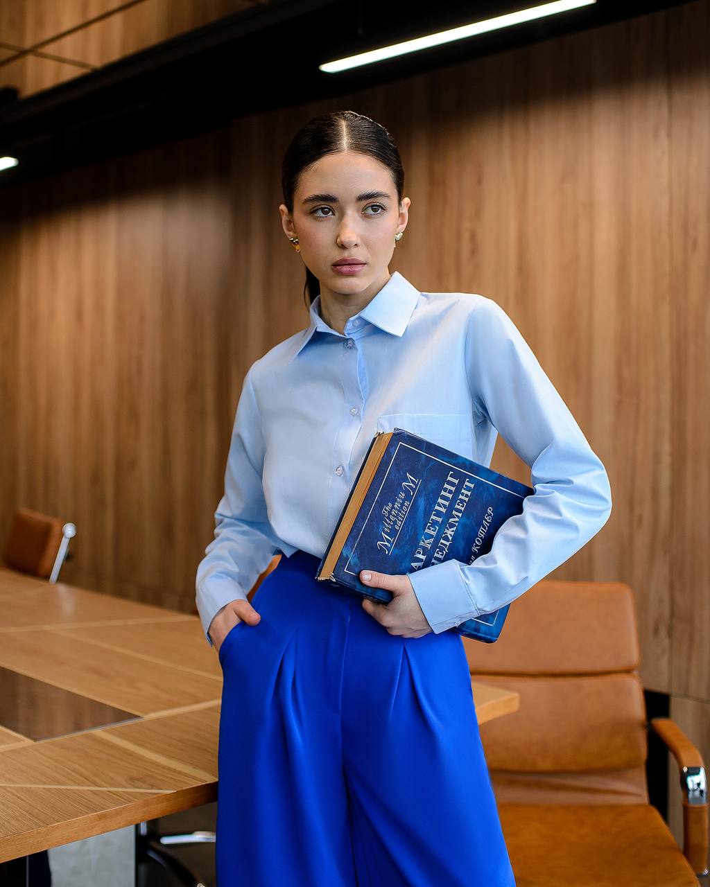 Woman in a light blue shirt and blue pants holding a book in an office setting.