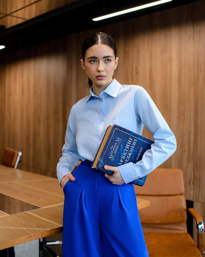 Woman in a light blue shirt and blue pants holding a book in an office setting.