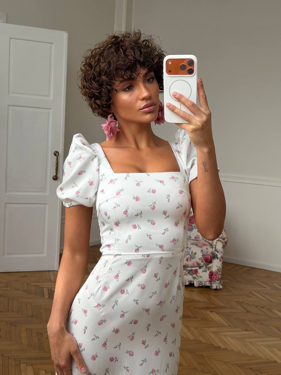 Woman taking a mirror selfie wearing a white floral dress in a room with wooden flooring.