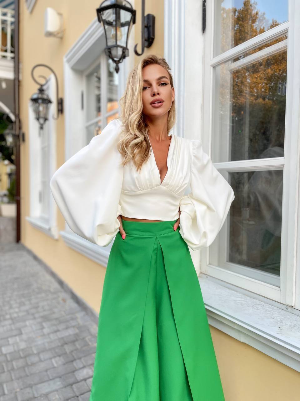 Woman in a white blouse and green pants standing in front of a building with a window.