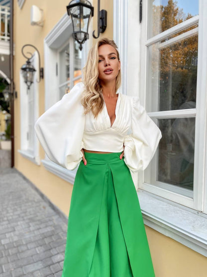 Woman in a white blouse and green pants standing in front of a building with a window.