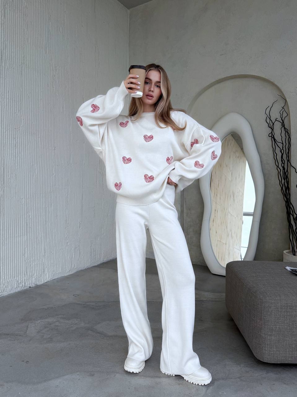 Person wearing a white sweatshirt with pink heart patterns and white pants in a modern interior setting.