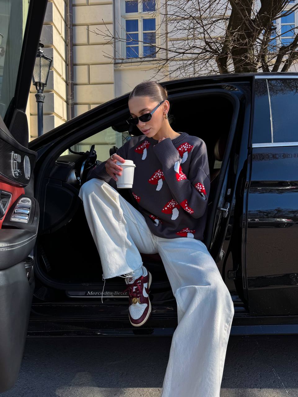 Person sitting in an open car door wearing a patterned sweater and white pants, holding a coffee cup.
