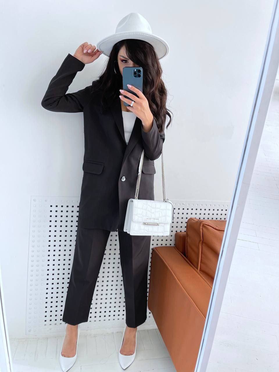 Person taking a mirror selfie wearing a black suit and white hat, holding a white handbag.