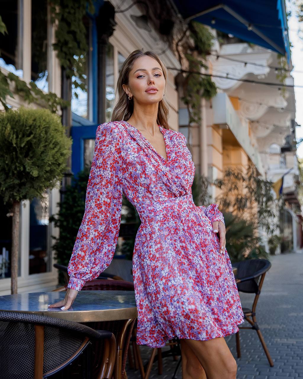 Woman in a floral dress standing outdoors near a building with plants.