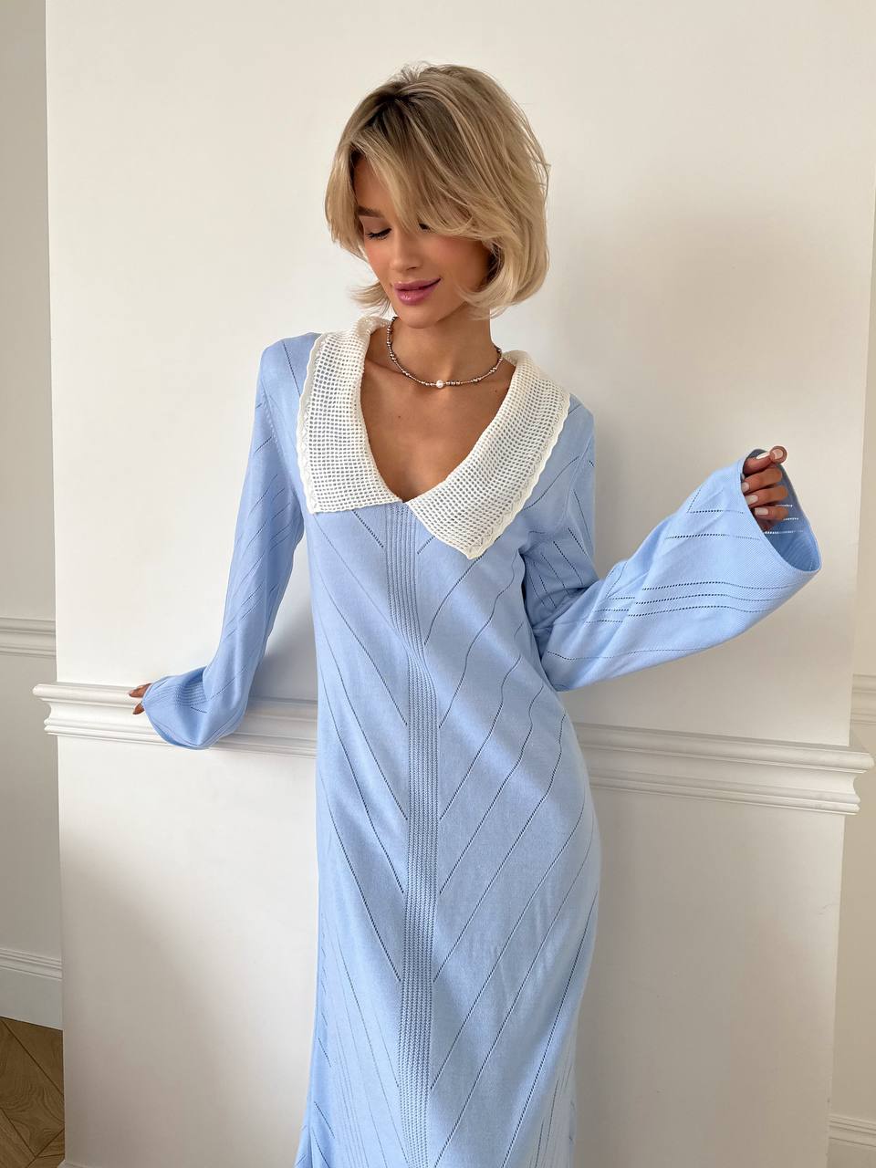 Woman wearing a light blue dress with a white collar against a white wall.