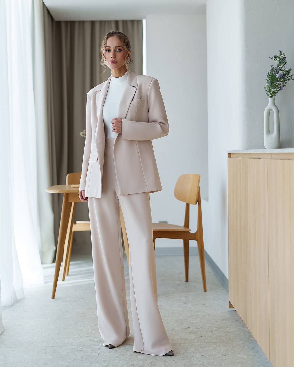 Woman in a beige suit standing in a modern room with wooden furniture and a vase.