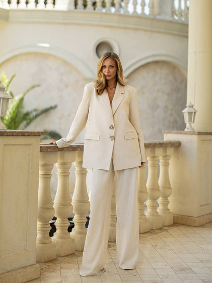 Woman in a beige suit standing on a balcony with decorative columns and plants.