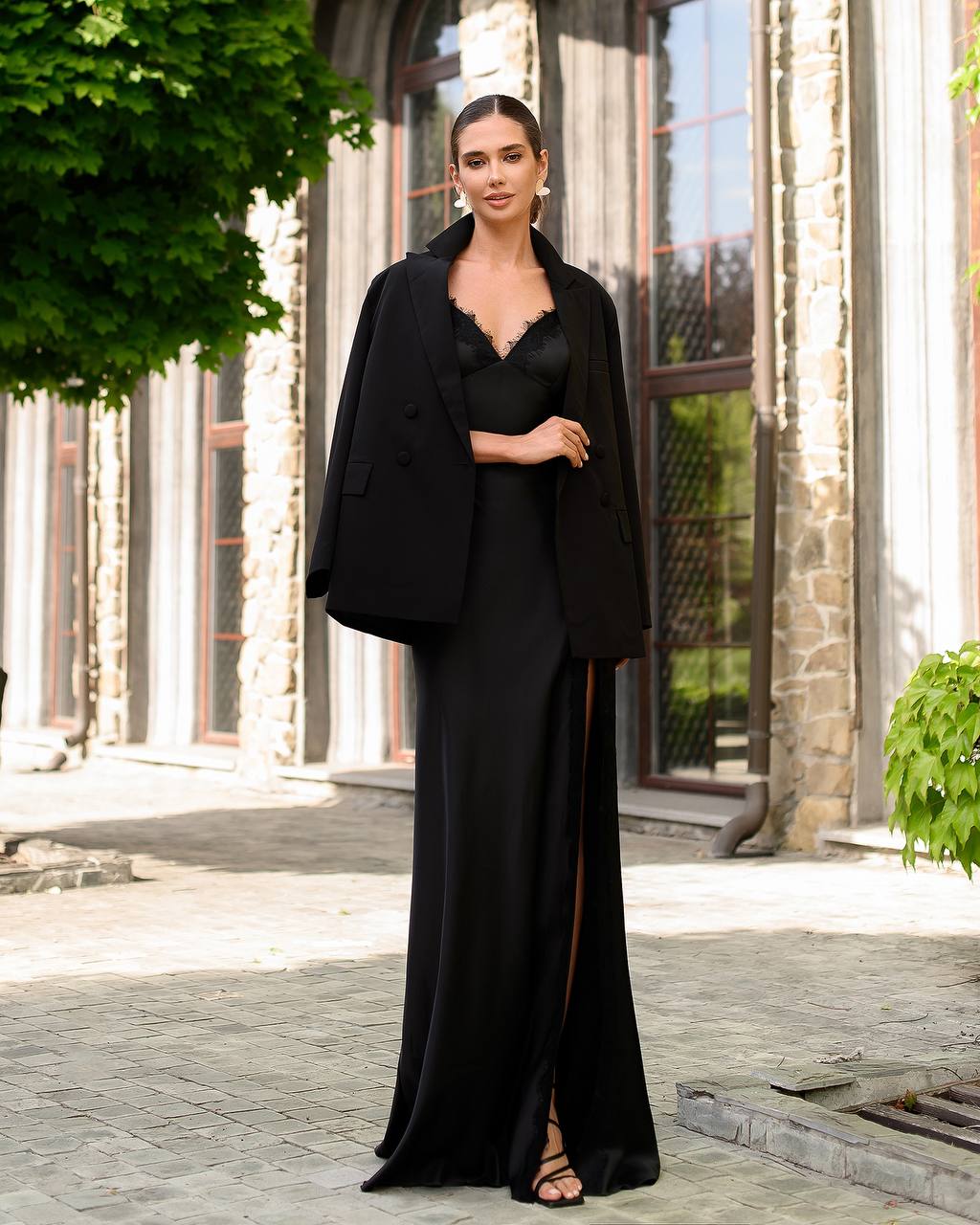 Woman in a black outfit standing outdoors with stone building and greenery in the background