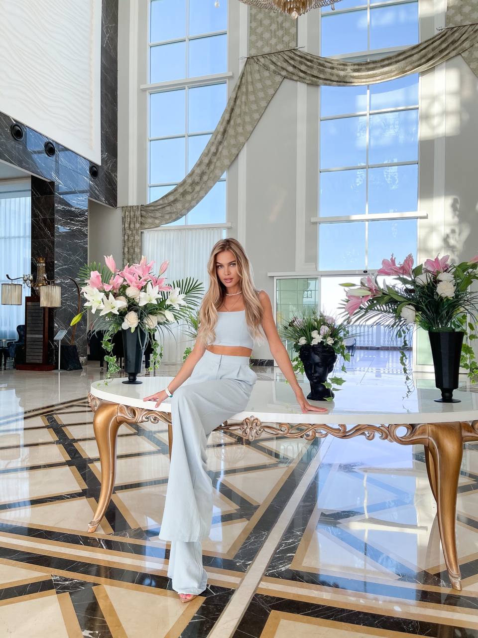 Woman in a white outfit sitting on a decorative chair in a luxurious lobby with large windows and floral arrangements.