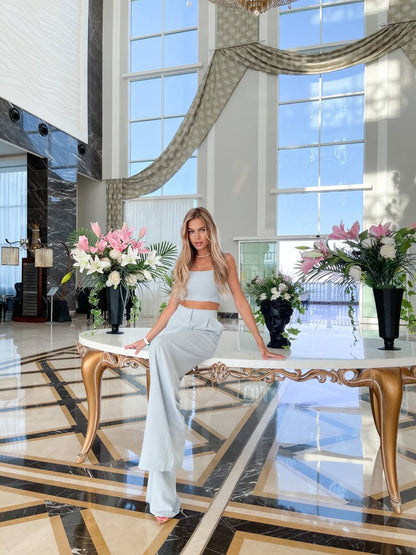 Woman in a white outfit sitting on a decorative chair in a luxurious lobby with large windows and floral arrangements.