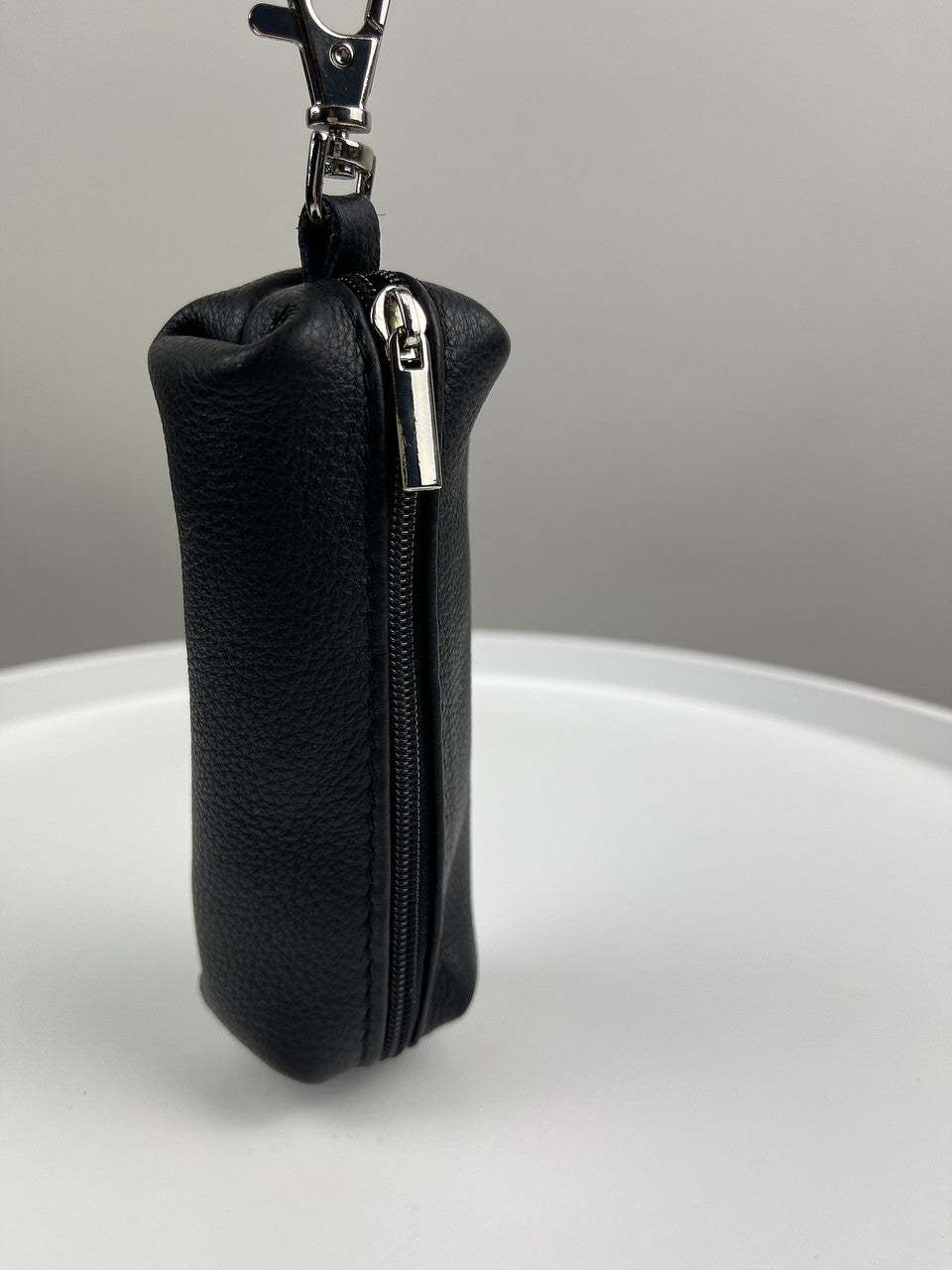 A small black pebbled leather zippered pouch with a silver metal zipper pull and a black keychain clasp at the top, standing upright on a white surface against a gray background.