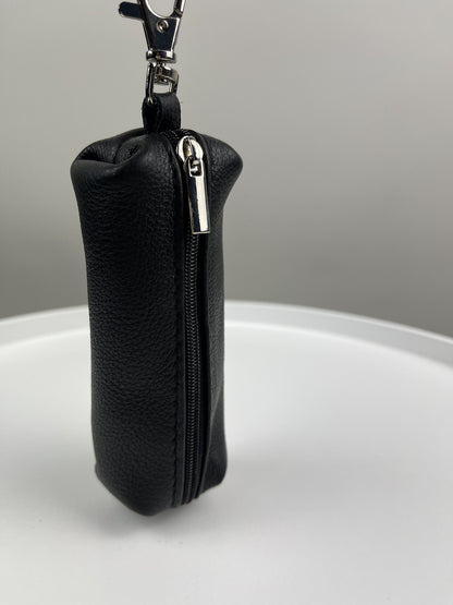 A small black pebbled leather zippered pouch with a silver metal zipper pull and a black keychain clasp at the top, standing upright on a white surface against a gray background.