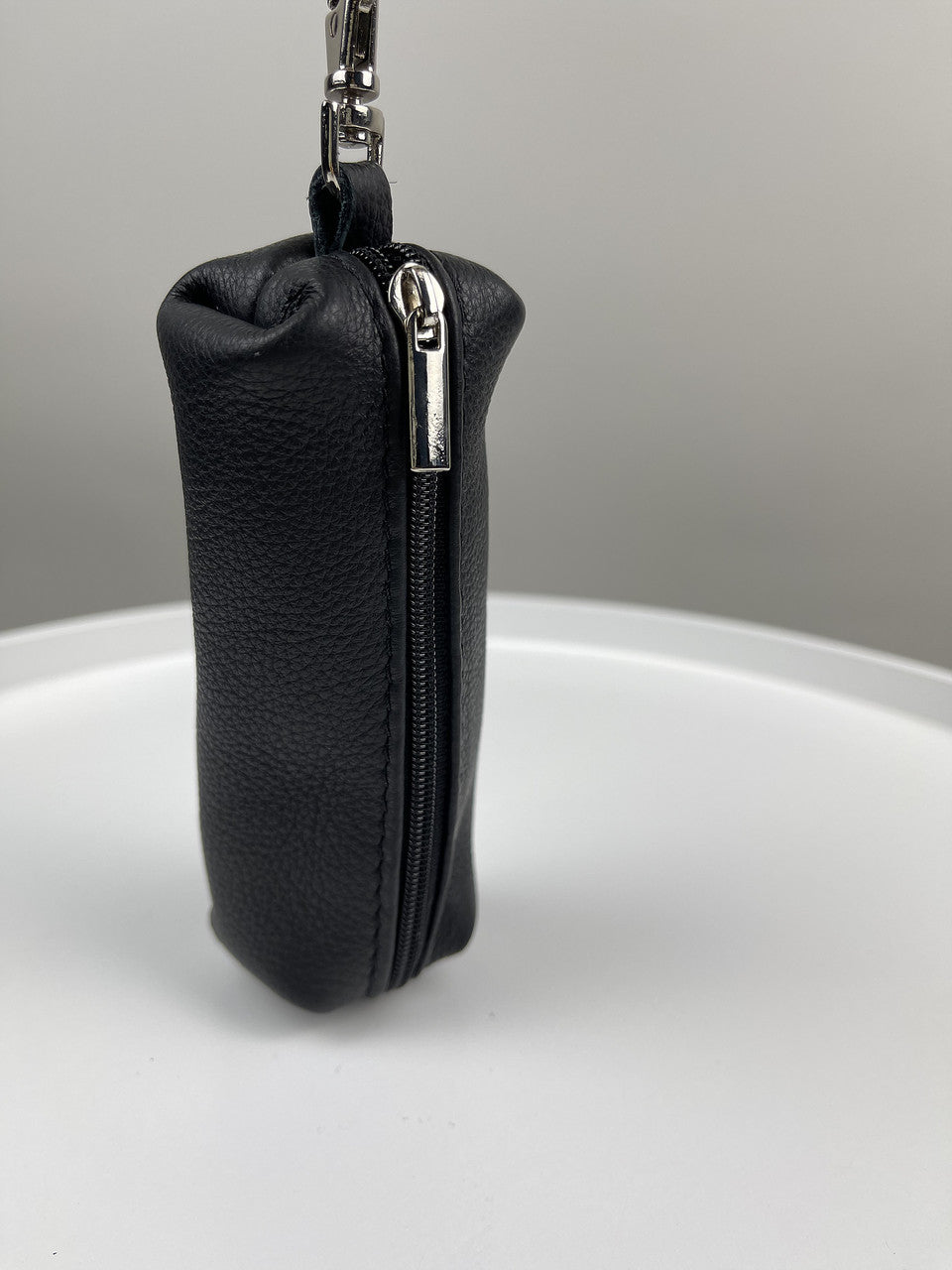 A small black textured leather zippered pouch with a silver zipper pull and attached metal keychain ring, standing upright on a white surface against a light gray background.