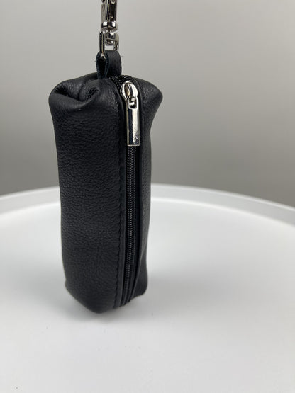 A small black textured leather zippered pouch with a silver zipper pull and attached metal keychain ring, standing upright on a white surface against a light gray background.