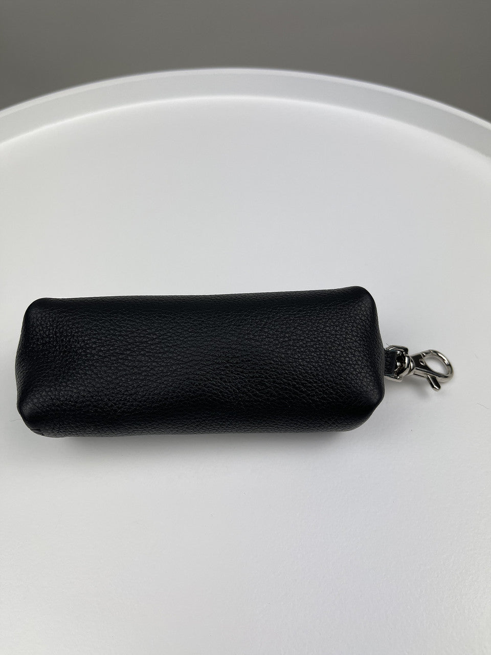 A small black pebbled leather pouch with a silver metal key ring clasp, resting on a white circular surface.