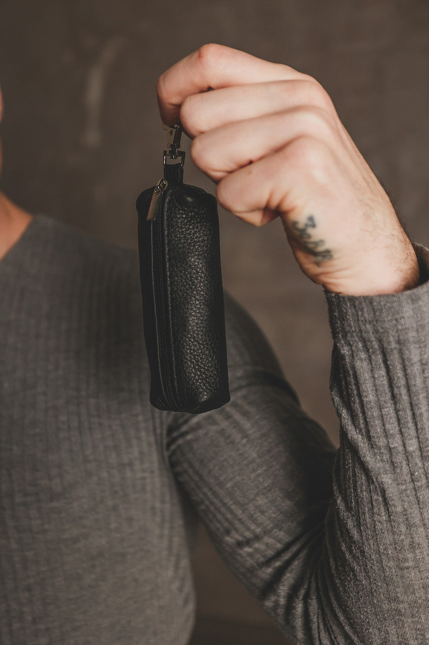 A close-up of a person's hand holding a small black pebbled leather pouch or case with a zipper and a small metal keyring, against a dark textured background.