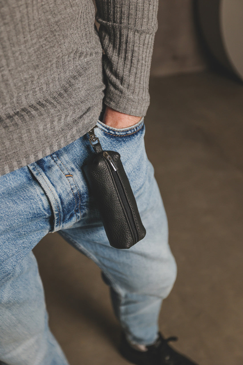 A close-up lifestyle shot of a person wearing a gray ribbed sweater, blue jeans, and black shoes, with a black textured leather zippered pouch hanging from their right pocket.