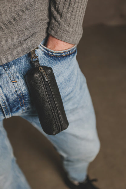 A close-up lifestyle shot of a person wearing a black pebbled leather zippered pouch clipped to their blue jeans, with a gray ribbed sweater visible.